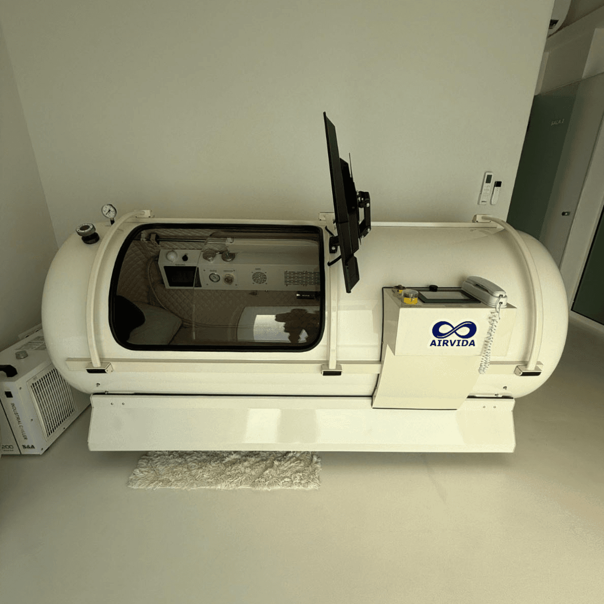AirVida Alpha 90 lying hard shell hyperbaric chamber in white with a long side window, a TV on an arm and an external control box
