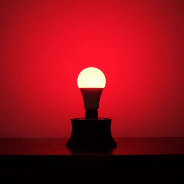 Chroma The Bulb, an A19 LED bulb standing in a black socket base on a wooden surface, the wall behind it washed in deep red Night Mode light, a still from the brand product animation