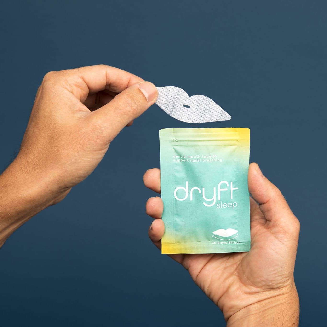 Hand holding a white lip-shaped Dryft Sleep mouth tape strip with a small centre slot above its teal and yellow pouch, on a plain navy background