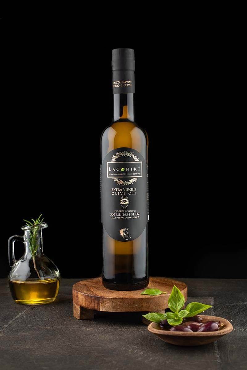 A 500 ml Laconiko Extra Virgin Olive Oil bottle with a black capsule and black oval label on a round wooden stand, beside a small glass cruet of oil holding a rosemary sprig and a wooden dish of dark olives with basil