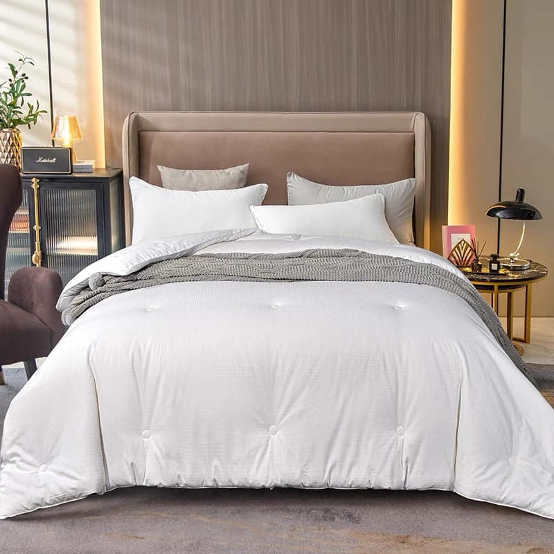 White tufted Lullabuy silk duvet spread over a made bed with two white pillows in front of two grey pillows at the headboard and a grey knit throw across the middle