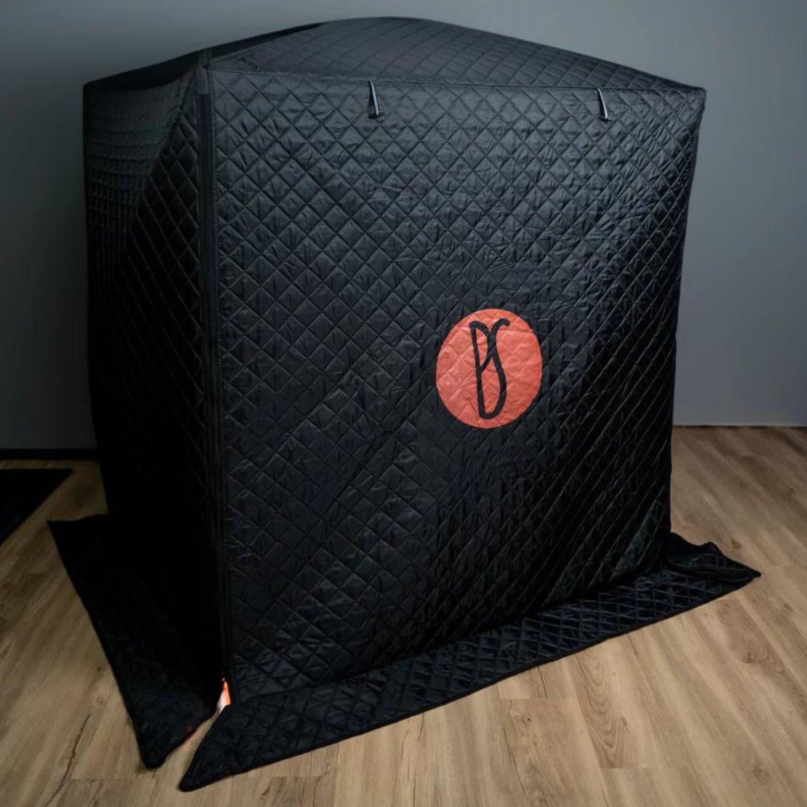 Prasanna infrared sauna pod tent standing closed on a wood floor, in black diamond quilted fabric with zip seams and a red circle logo on the front panel