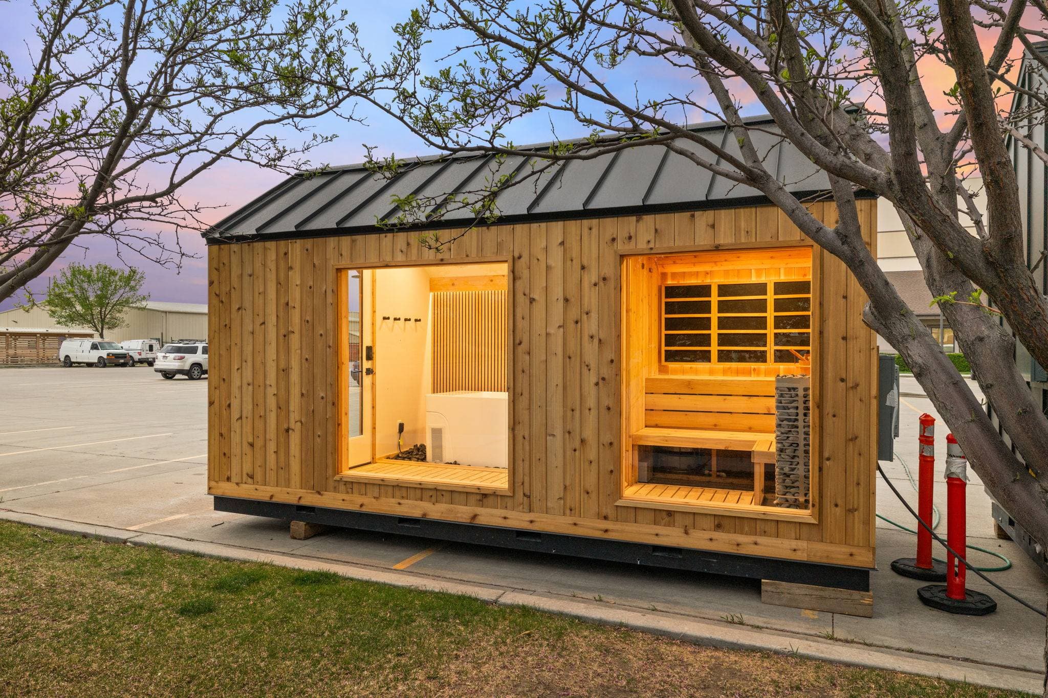 Sweaty Yeti Sauna Plunge 8 by 17 foot modular building at dusk, shown with optional cedar siding and a metal roof, with the white cold plunge tub visible through the left window and the cedar sauna with its stone tower heater through the right window