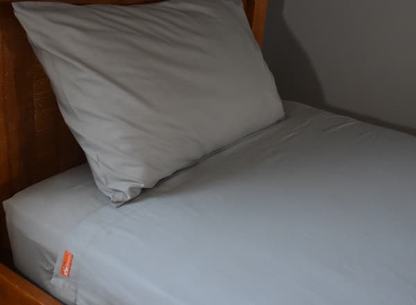 Grey ZipSheets cotton sheet set on a single bed with a woven wooden headboard, the top sheet laid flat over the fitted sheet, one grey pillowcase and a small orange ZipSheets label on the side of the sheet
